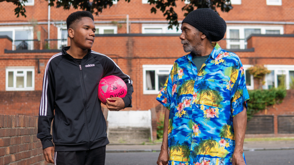 An older man and a young man with a football walking down the street talking.