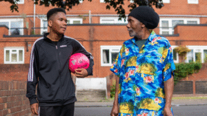 An older man and a young man with a football walking down the street talking.