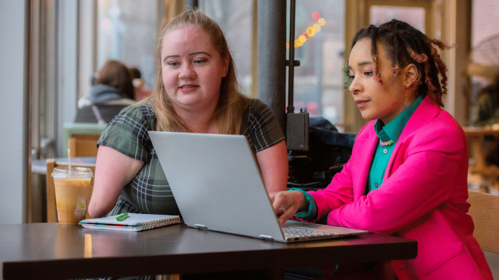 Two people in bright clothing sat in a cafe working or studying or learning with a laptop and pen and notebook.