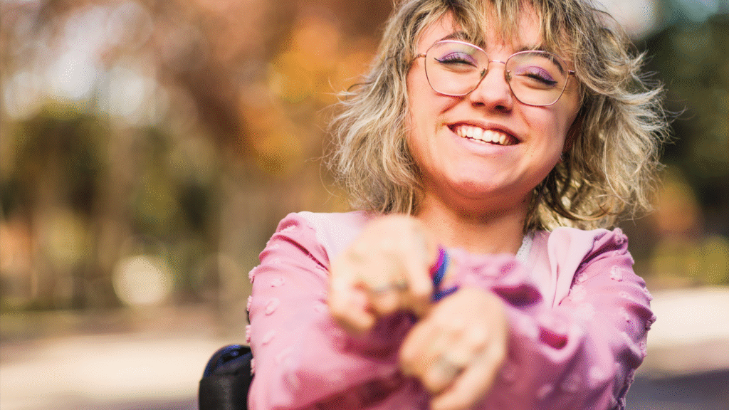 A woman with glasses and a pink blouse smiling outside with her hands forwards towards to the camera.